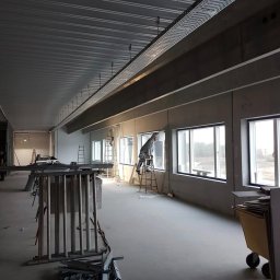 MADO Mateusz Dowejko - Wnętrze biura w trakcie wykańczania: pracownik na drabinie przy oknie, widoczne instalacje pod sufitem, elementy konstrukcyjne i narzędzia na podłodze.