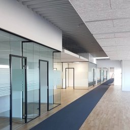 MADO Mateusz Dowejko - Nowoczesne biuro z przeszklonymi ścianami działowymi, drewnianą podłogą i szarym dywanem w korytarzu. Widoczne detale wykończenia sufitu i oświetlenia.