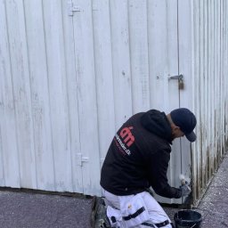 MADO Mateusz Dowejko - Mężczyzna w czarnej bluzie z logo maluje białą, drewnianą ścianę pędzlem. Obok stoi czarne wiadro z farbą. Widoczne ślady starej farby i świeżo pomalowany fragment.