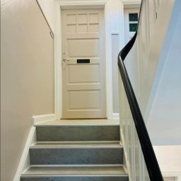 MADO Mateusz Dowejko - Klatka schodowa po remoncie: szare stopnie, białe barierki i beżowe ściany. Drzwi wejściowe w jasnym odcieniu z czarną skrzynką na listy. Widok z dołu schodów.