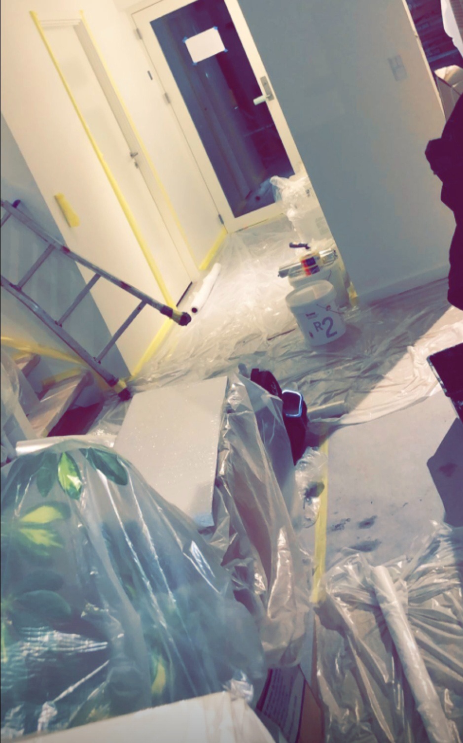 Przygotowania do malowania: zabezpieczona folią podłoga, ściany oklejone taśmą malarską, drabina i wiadro z farbą w pomieszczeniu. Widoczny fragment rośliny.