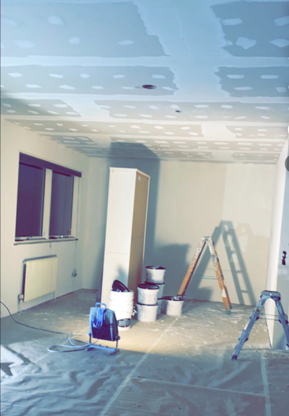 Pokój w trakcie szpachlowania sufitu, widoczne drabiny, wiaderka z masą szpachlową, folia ochronna na podłodze i lampa budowlana. Etap prac wykończeniowych.