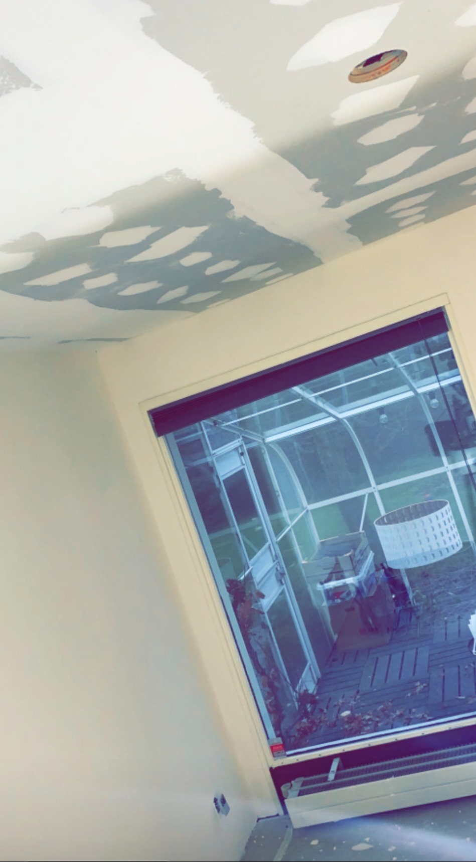 Szpachlowany sufit z widocznymi łatami gładzi, przygotowany do malowania. W tle okno z widokiem na ogród zimowy.