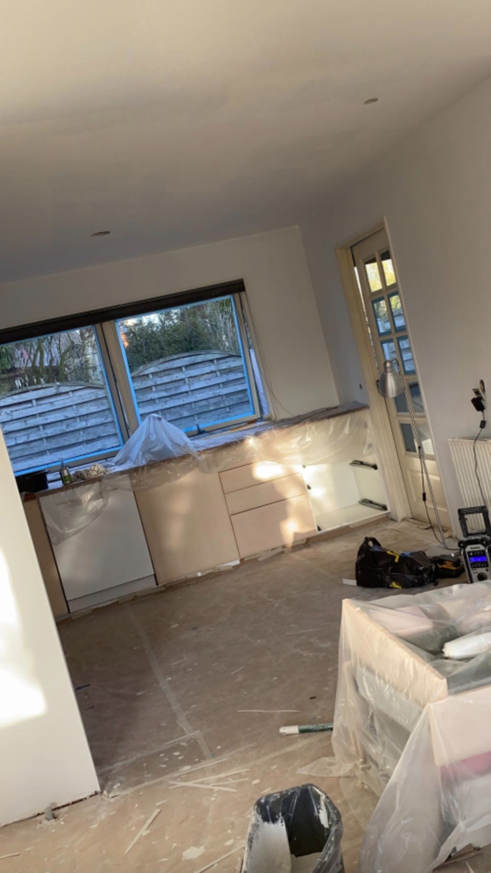 Remont kuchni w trakcie: zabezpieczone folią meble, okno i podłoga. Widoczne narzędzia malarskie i puszka z farbą. Ściany przygotowane do malowania.