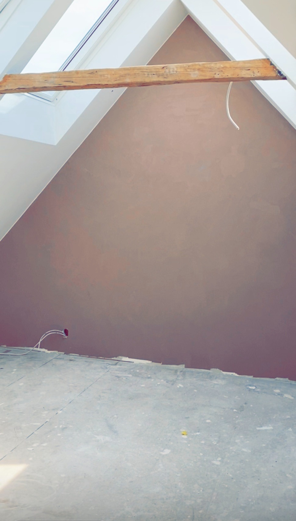 Poddasze ze ścianą pokrytą świeżą, brązową gładzią. Widoczne okno dachowe z drewnianą belką i wystające kable elektryczne. Podłoga przygotowana do dalszych prac wykończeniowych.