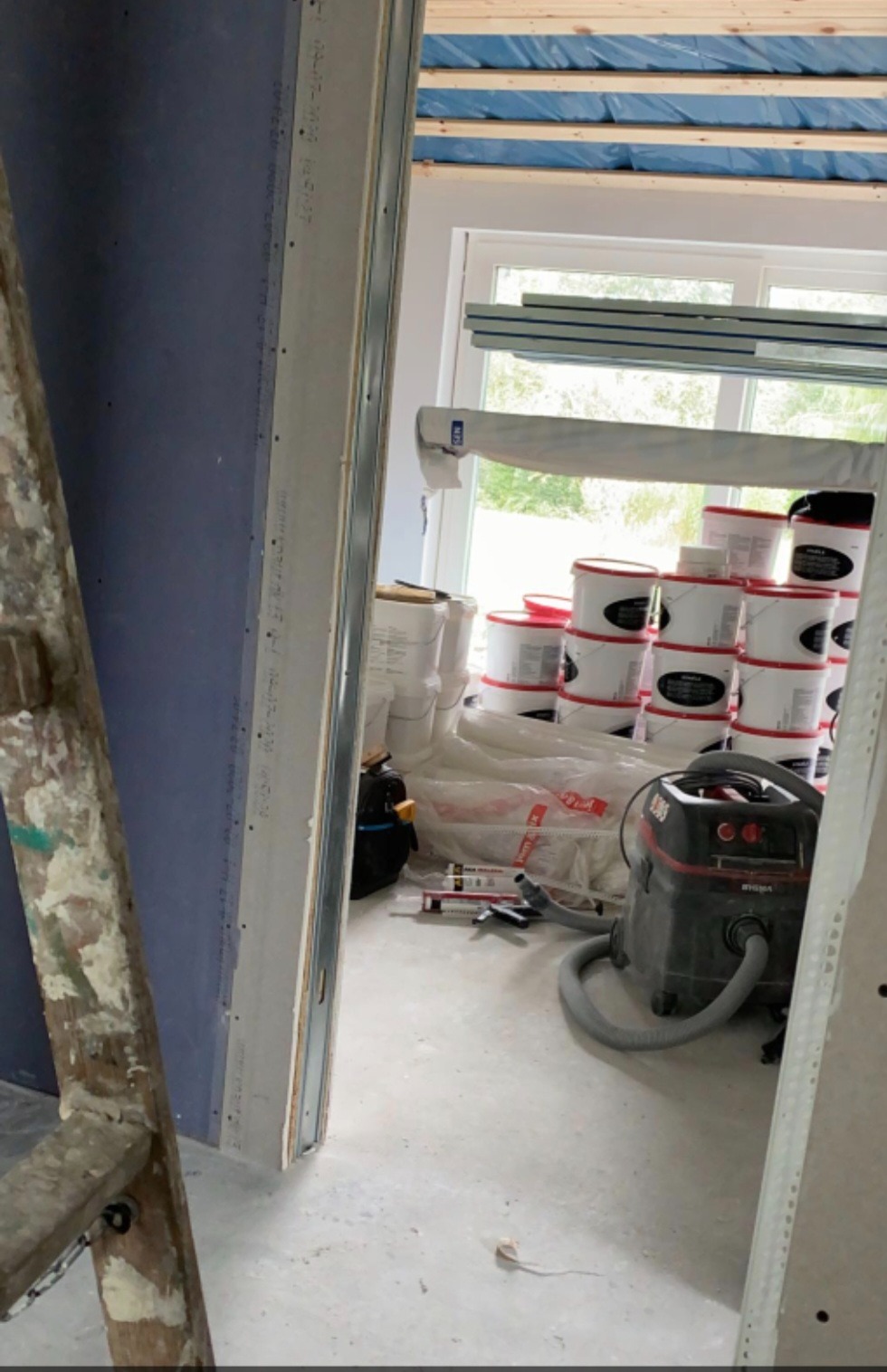 Wnętrze w trakcie prac wykończeniowych: ściana z płyt gipsowych, drabina malarska, wiadro z farbą, odkurzacz przemysłowy. Widoczne elementy konstrukcji sufitu.