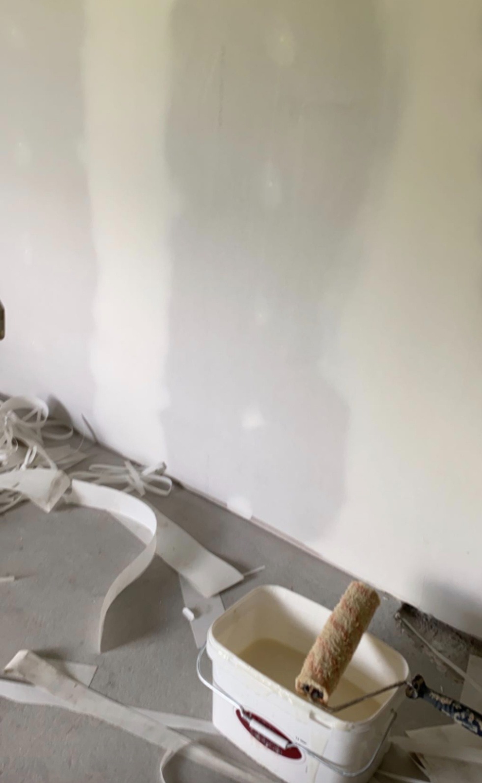 Ściana po szpachlowaniu, gotowa do malowania. Widoczne narzędzia: wałek w wiadrze z farbą i taśmy malarskie na podłodze. Przygotowanie do malowania ścian.