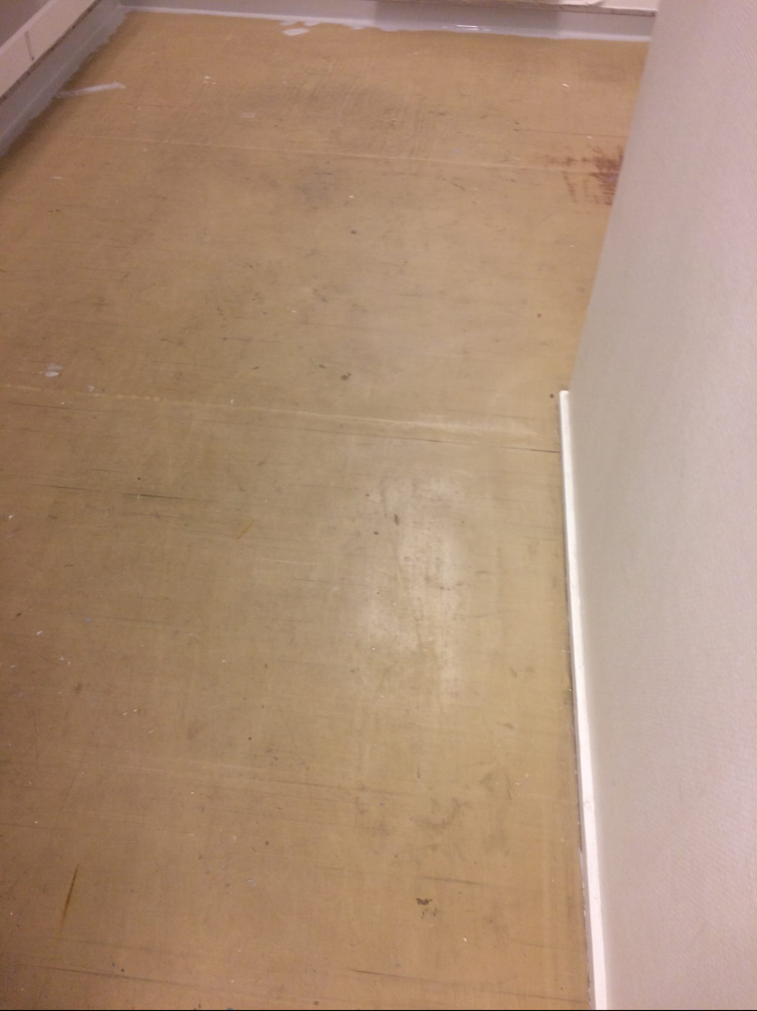 Brązowa, stara podłoga z widocznymi śladami użytkowania, przylegająca do białej ściany. Widoczne drobne zabrudzenia i rysy na powierzchni podłogi. Ujęcie z góry.