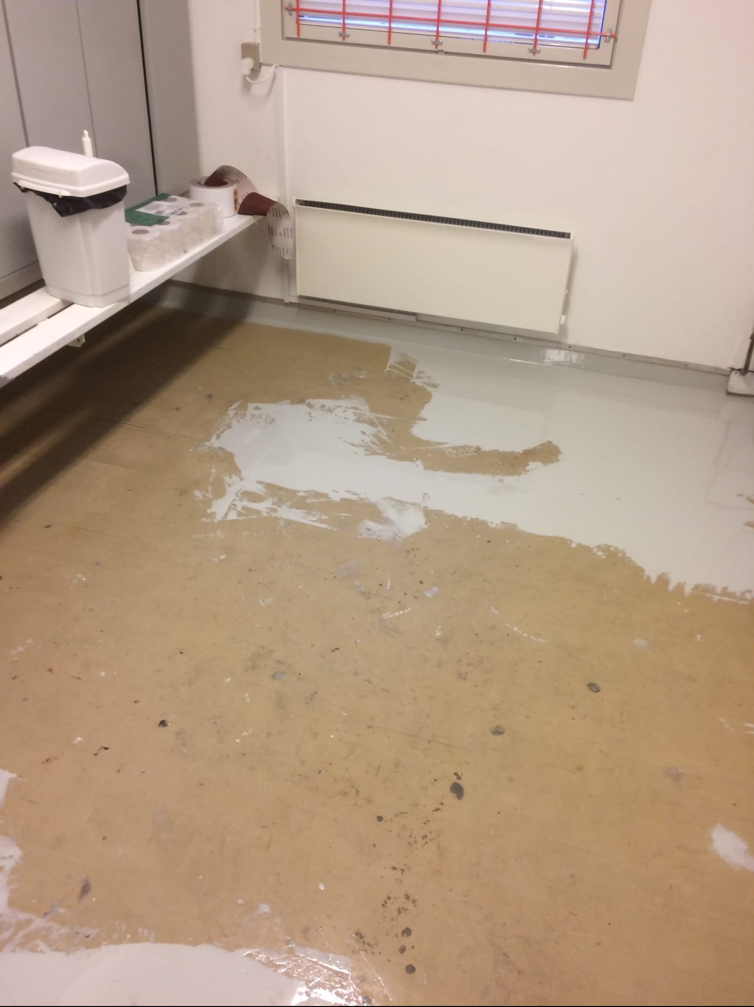 Wnętrze w trakcie wylewania posadzki żywicznej. Widoczna częściowo pokryta podłoga, biały grzejnik i akcesoria malarskie na półce. Etap prac wykończeniowych.