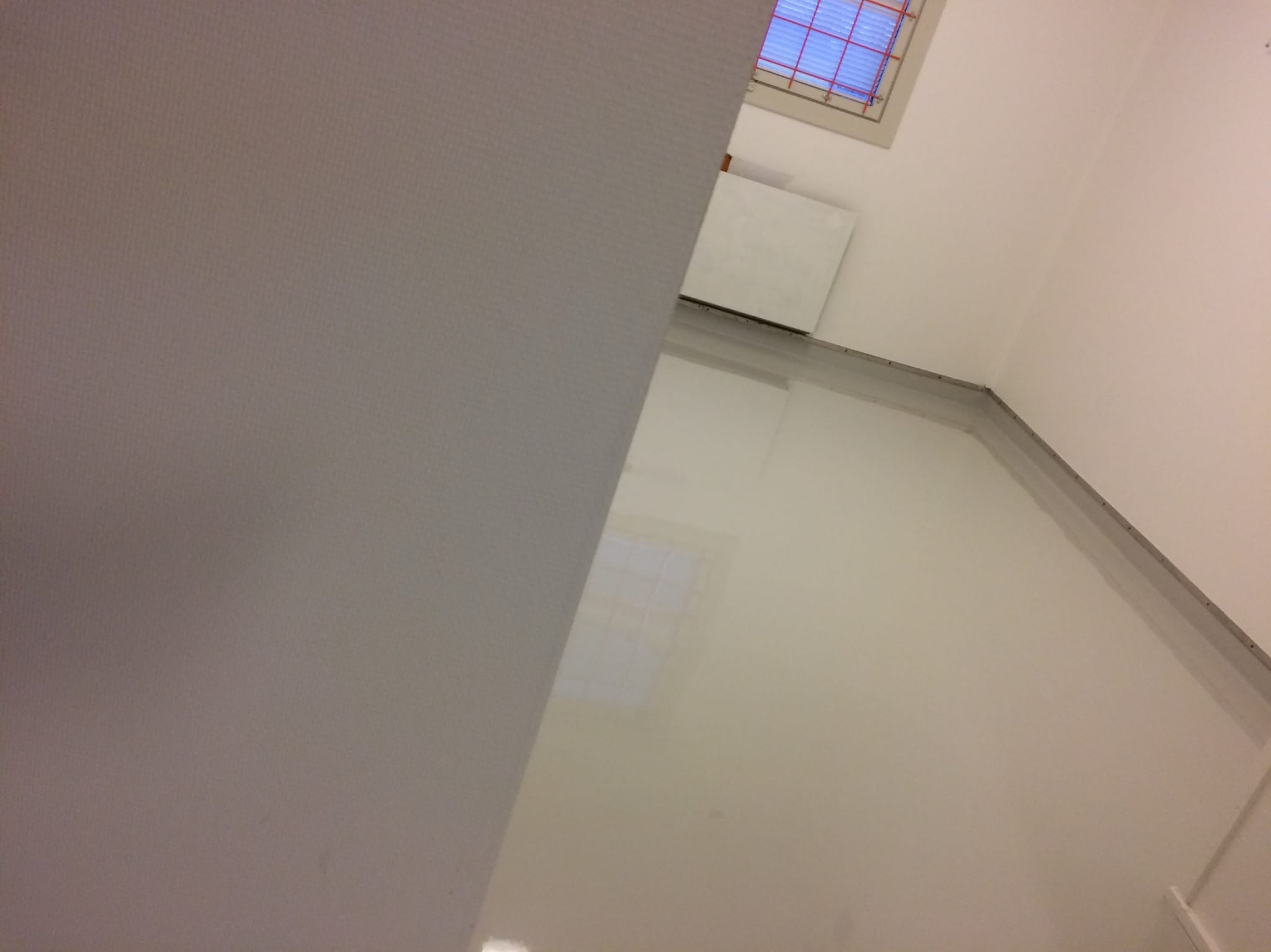 Błyszcząca, jasna posadzka żywiczna w pokoju z białymi ścianami i grzejnikiem pod oknem. Minimalistyczne wykończenie, nowoczesny design.