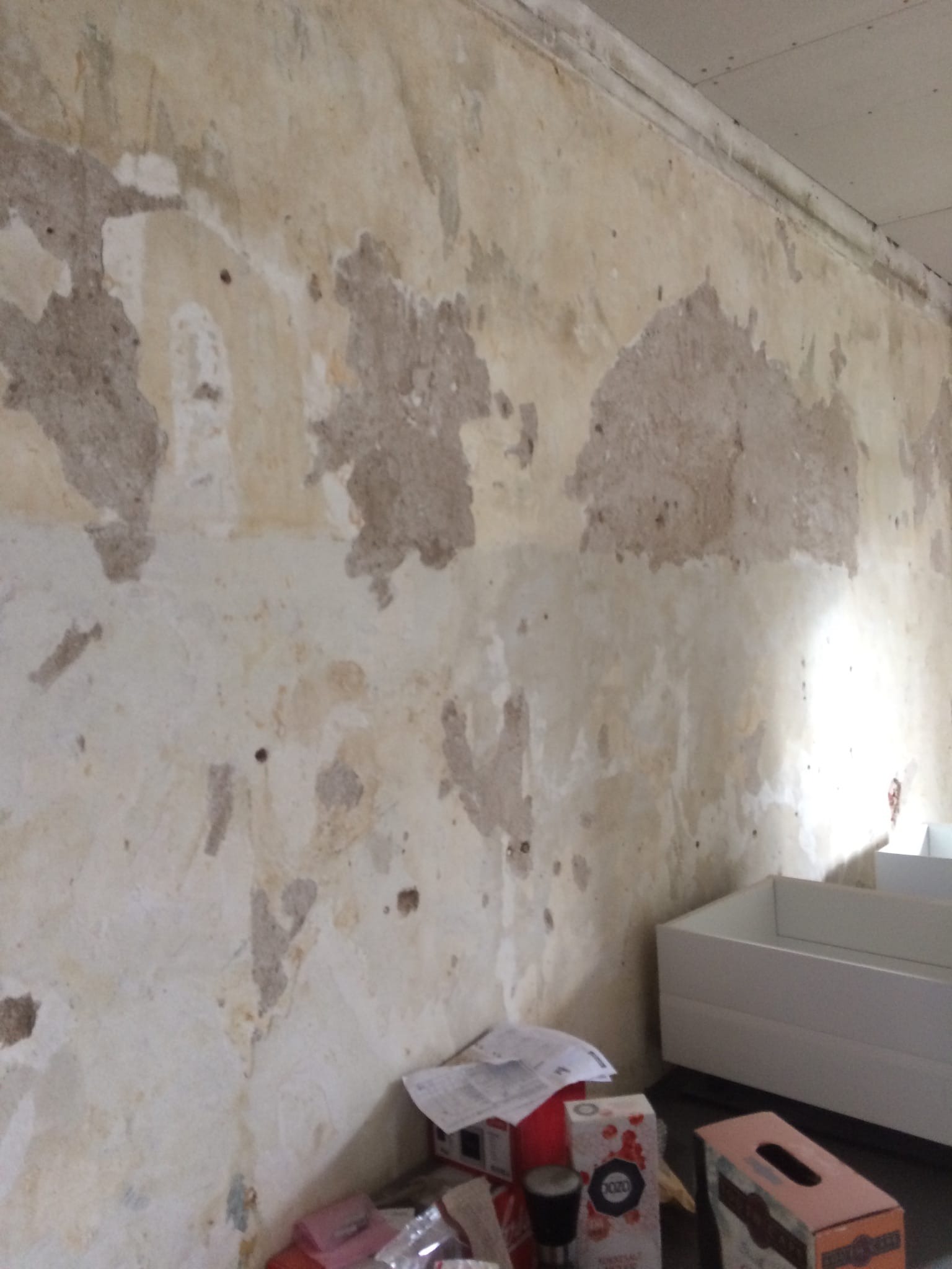 Nierówna, stara ściana z ubytkami tynku, przygotowana do remontu. Widoczne fragmenty starej farby i pozostałości po usuniętym tynku. W tle białe pudło i drobne przedmioty.
