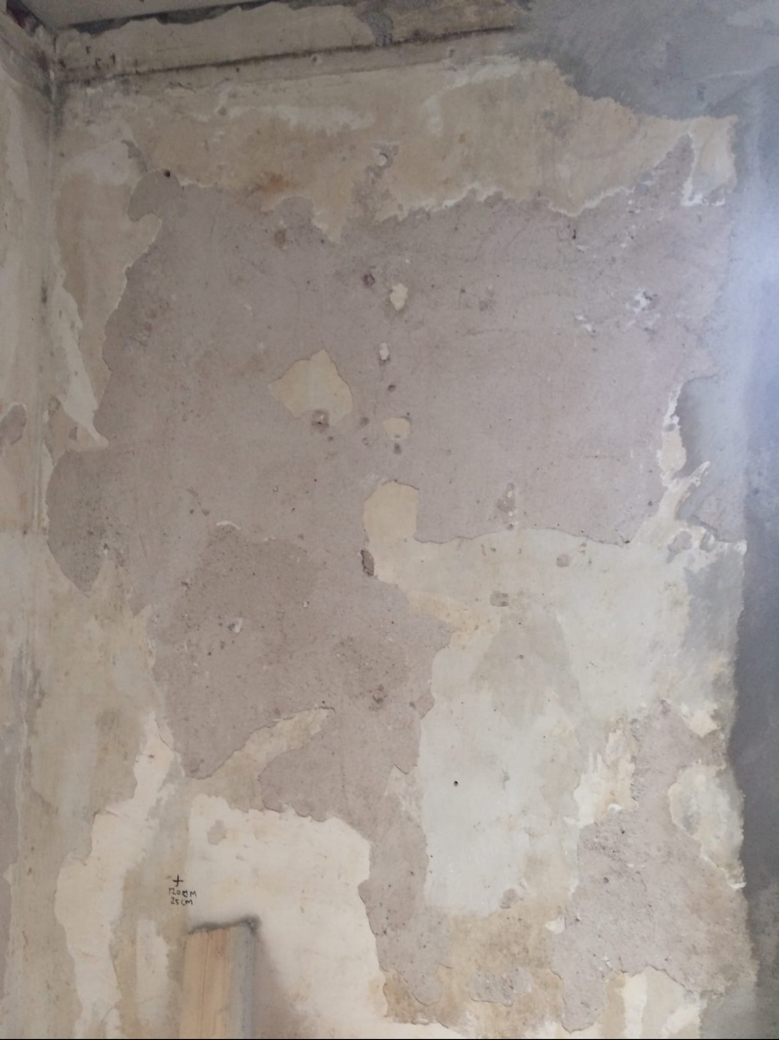 Narożnik pokoju z nierówną, przygotowaną do szpachlowania ścianą. Widoczne ślady po starych powłokach i notatki ołówkiem na ścianie. Fragment drewnianej listwy przypodłogowej.
