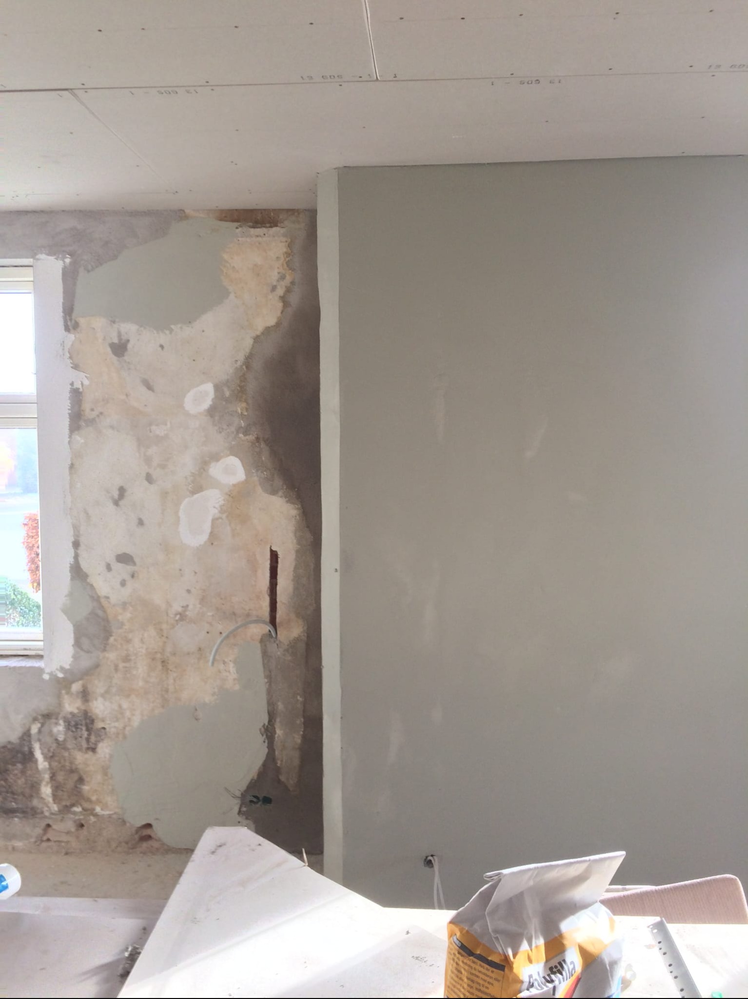 Ściana w trakcie szpachlowania: fragment surowej ściany obok gładkiej, wyszpachlowanej powierzchni. Widoczne narzędzia i worek z zaprawą. Ujęcie z bliska.