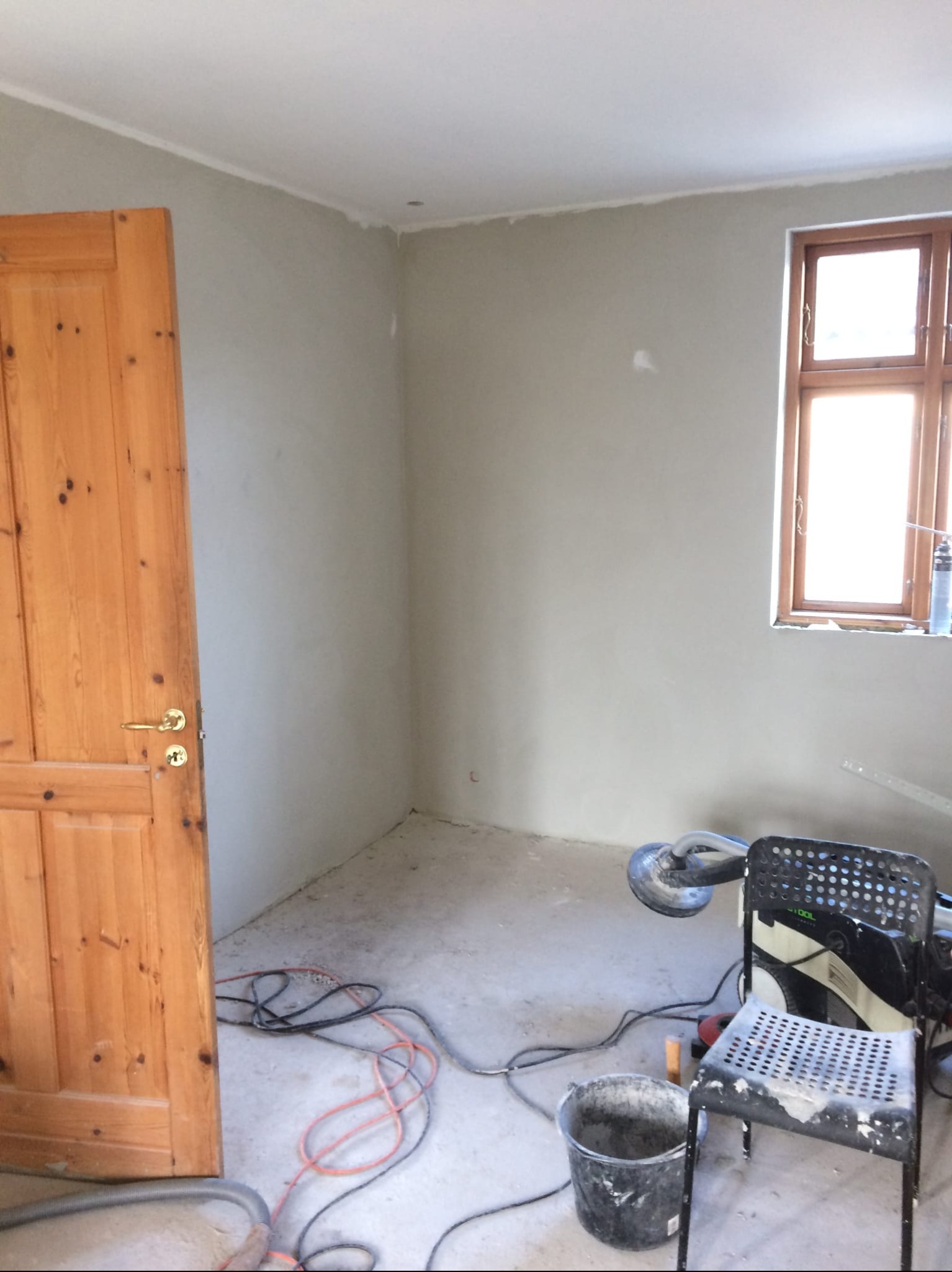 Pokój po szpachlowaniu ścian, widoczne narzędzia: szlifierka, wiadro z gładzią, krzesło. Drewniane drzwi po lewej, okno z drewnianą ramą po prawej.