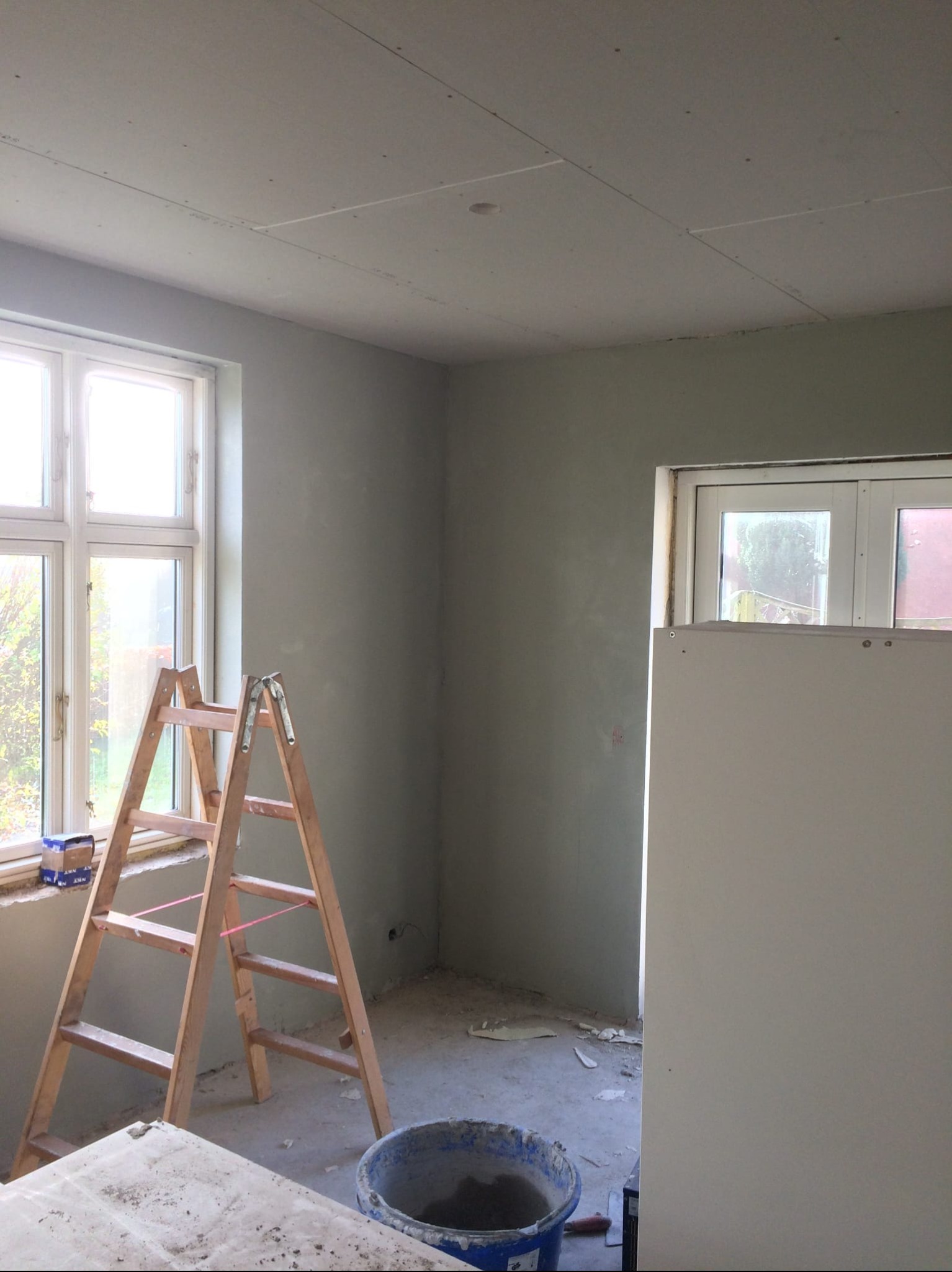 Pokój w trakcie szpachlowania: drewniana drabina przy oknie, ściany pokryte szpachlą, sufit z płyt karton-gips, wiadro z zaprawą na podłodze. Widoczne niedoskonałości.