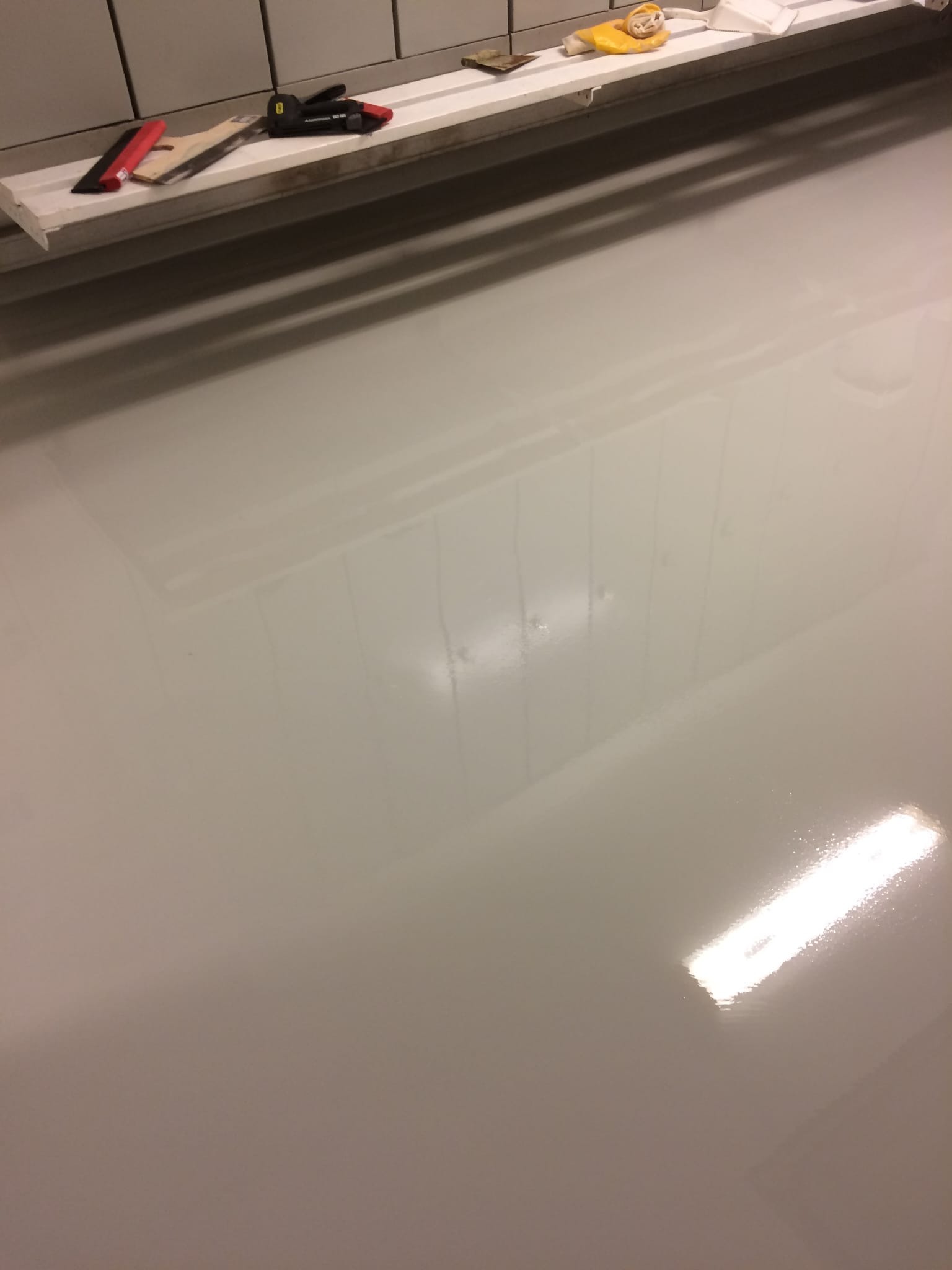 Jasnoszara, gładka posadzka żywiczna z odbiciem światła, narzędzia na półce w tle. Minimalistyczne wykończenie podłogi w neutralnej tonacji.