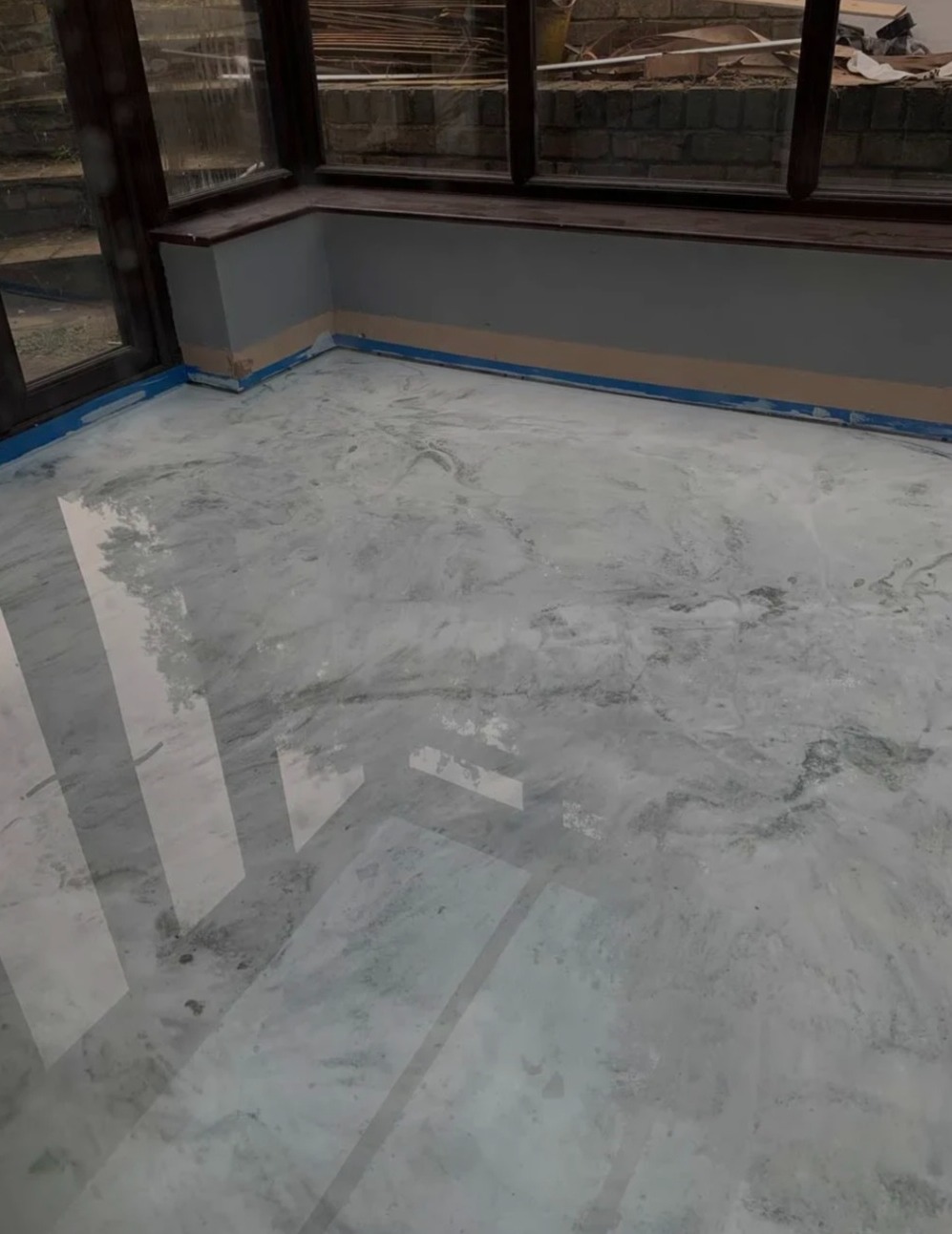 Błyszcząca, jasnoszara posadzka żywiczna z efektem marmuru w przeszklonym pomieszczeniu. Widoczne odbicia światła i taśmy malarskie przy ścianach.