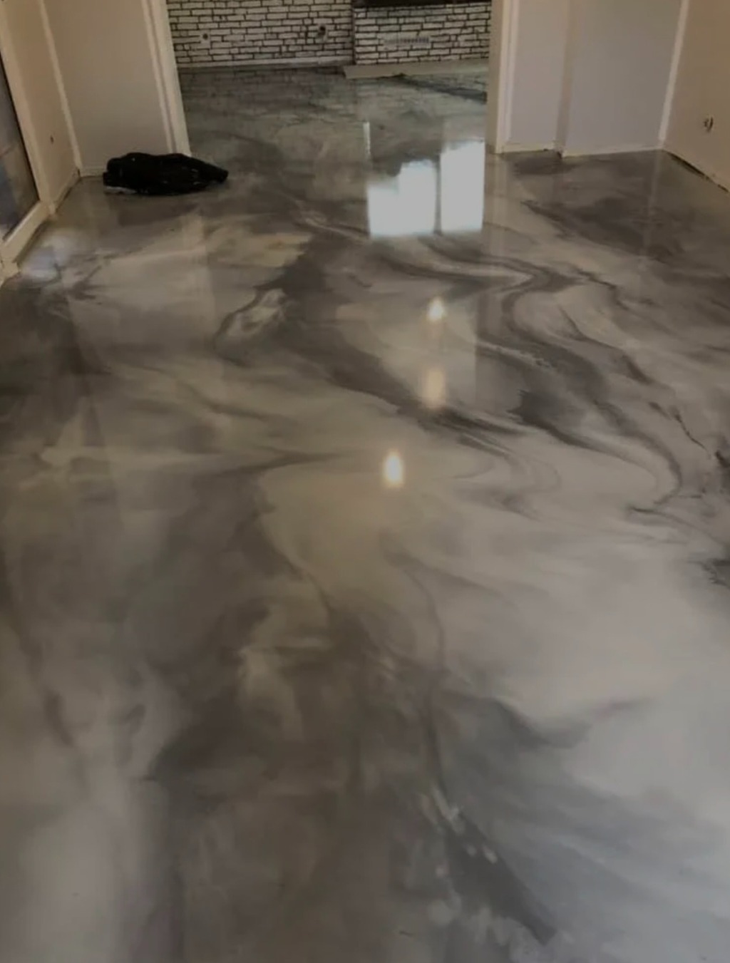 Błyszcząca, szara posadzka żywiczna z efektem marmuru, odbijająca światło okien. W tle widoczny fragment ściany z cegły i przejścia do innych pomieszczeń.