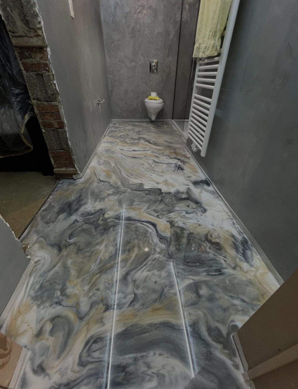 Błyszcząca, marmurkowa posadzka żywiczna w łazience z szarą ścianą i białym grzejnikiem drabinkowym. Widok z perspektywy wejścia, z fragmentem surowej cegły.