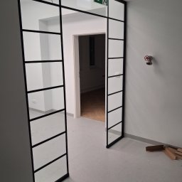 Usługi remontowo-budowlane Marek Tomczyk - Nowoczesne, przeszklone drzwi z czarną ramą w biurze, widok na korytarz z parkietem. Nad drzwiami znak ewakuacyjny. Na podłodze kartony i narzędzia.