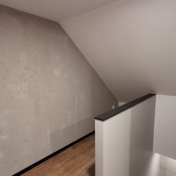 Usługi remontowo-budowlane Marek Tomczyk - Wykończone poddasze ze schodami: drewniana podłoga, szara ściana z efektem betonu, białe ściany i sufit. Minimalistyczny design z czarnymi akcentami.