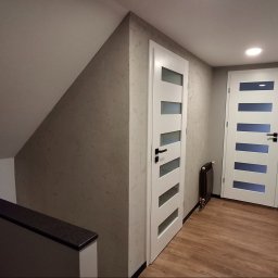 Usługi remontowo-budowlane Marek Tomczyk - Korytarz na poddaszu z dwiema białymi, przeszklonymi drzwiami i drewnopodobną podłogą. Ściany w odcieniach szarości, jedna ze strukturą. Widoczny fragment schodów.