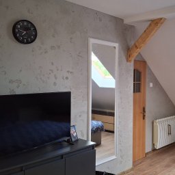 Usługi remontowo-budowlane Marek Tomczyk - Pokój na poddaszu z szarą ścianą o fakturze betonu, czarnym zegarem i telewizorem. W lustrze odbija się fragment sypialni. Widoczna drewniana belka i drzwi.