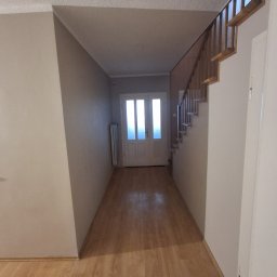 Usługi remontowo-budowlane Marek Tomczyk - Jasne wnętrze przedpokoju z drewnianą podłogą, schodami i białymi drzwiami wejściowymi. Widoczny grzejnik i neutralne kolory ścian.