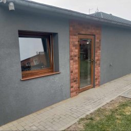 Usługi remontowo-budowlane Marek Tomczyk - Szary budynek z oknem i drzwiami obramowanymi cegłą, widok z zewnątrz, kostka brukowa, trawa. Elewacja o fakturze baranka. Okno z drewnianą ramą.