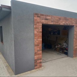 Usługi remontowo-budowlane Marek Tomczyk - Szary garaż z ceglanym obramowaniem wjazdu, widok z zewnątrz. Wnętrze garażu częściowo widoczne, z widocznymi narzędziami i przedmiotami.