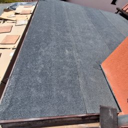 Usługi remontowo-budowlane Marek Tomczyk - Szczegółowe ujęcie dachu pokrytego papą termozgrzewalną, z widocznymi elementami wykończeniowymi i fragmentem ściany budynku w ciepłym, pomarańczowym odcieniu.