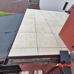 Usługi remontowo-budowlane Marek Tomczyk - Ułożone płytki na zewnątrz budynku, widok z góry, fragment dachu i ściany. Widoczne rusztowanie i elementy konstrukcyjne. Jasne płytki kontrastują z ciemnym dachem.