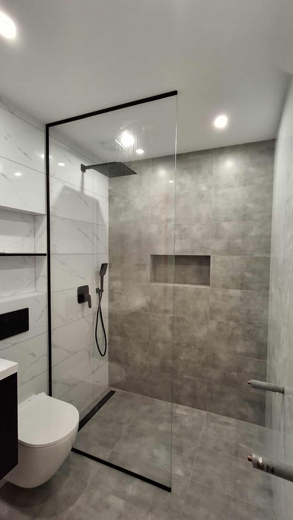 Nowoczesna łazienka z kabiną prysznicową typu walk-in, wykończona szarymi i białymi płytkami. Widoczna toaleta podwieszana i czarna armatura.