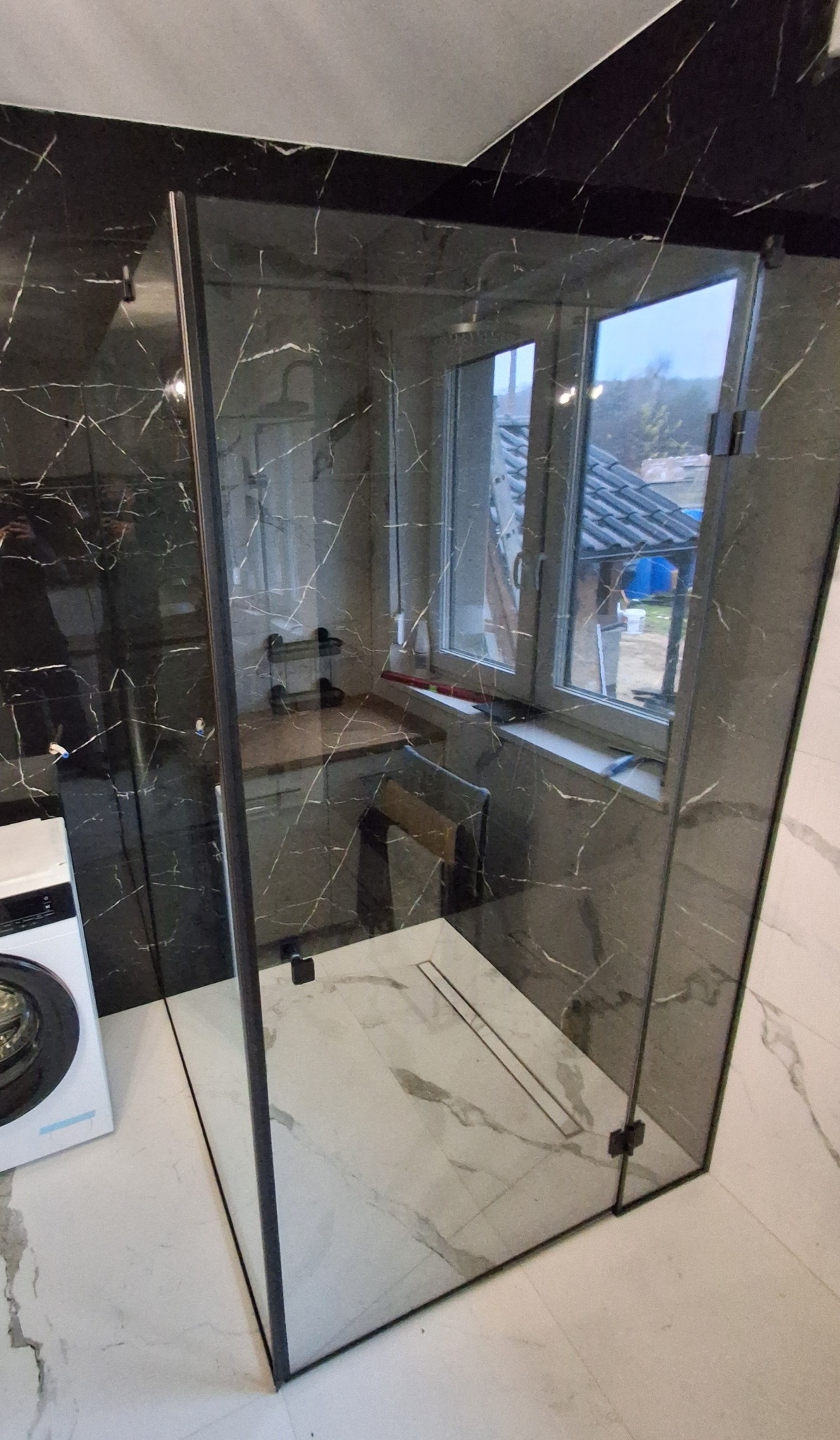 Nowoczesna kabina prysznicowa z czarnymi profilami i marmurowymi płytkami w łazience. Widoczna pralka i okno z widokiem na zewnątrz.