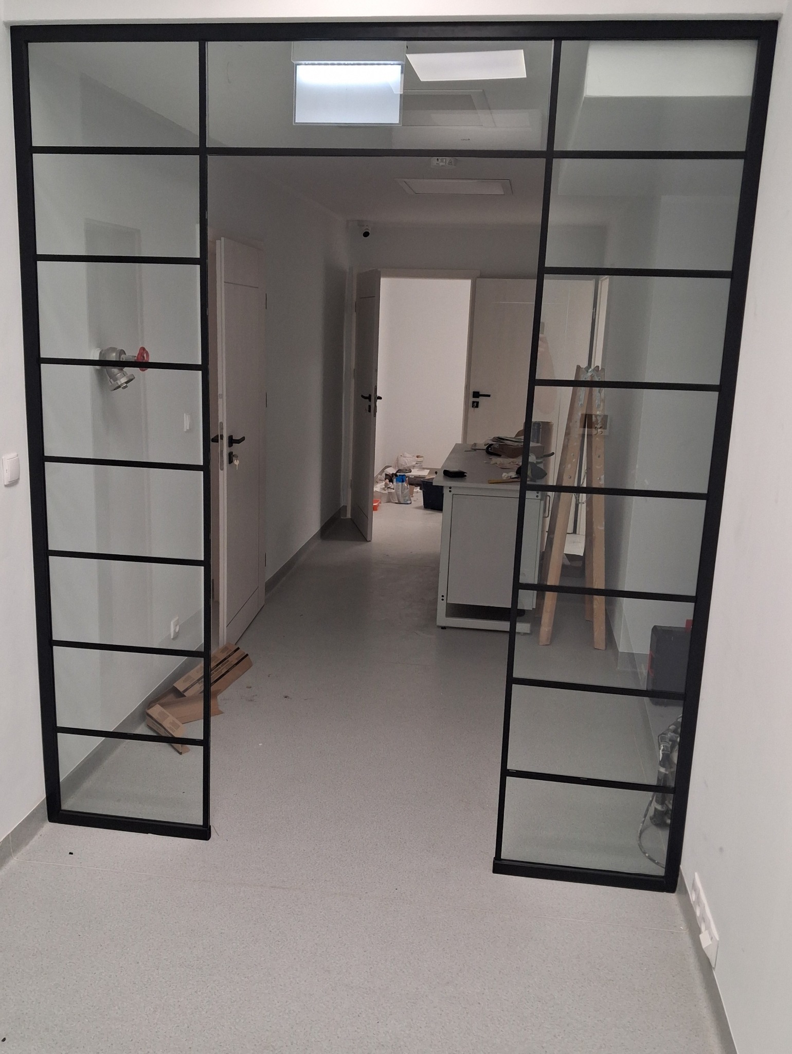 Nowoczesne, szklane drzwi przesuwne z czarnymi szprosami oddzielają pomieszczenia biurowe. Widoczny fragment wnętrza z biurkiem i schodami.