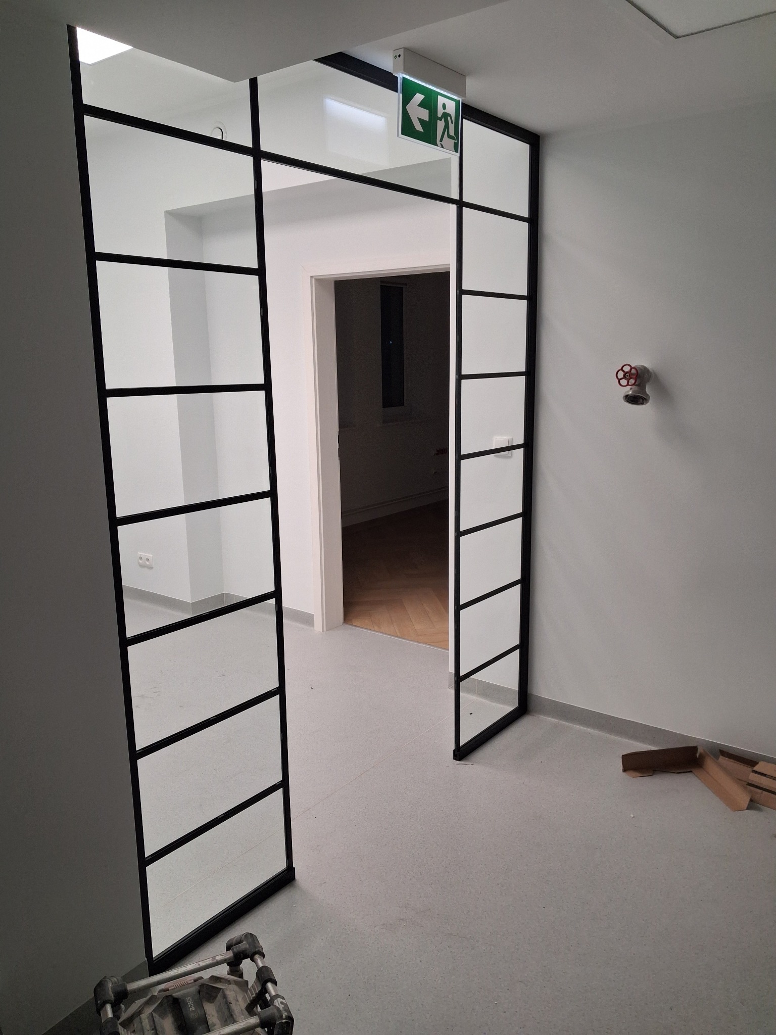 Nowoczesne, przeszklone drzwi z czarną ramą w biurze, widok na korytarz z parkietem. Nad drzwiami znak ewakuacyjny. Na podłodze kartony i narzędzia.