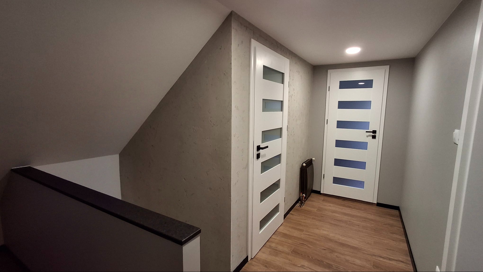 Korytarz na poddaszu z dwiema białymi, przeszklonymi drzwiami i drewnopodobną podłogą. Ściany w odcieniach szarości, jedna ze strukturą. Widoczny fragment schodów.