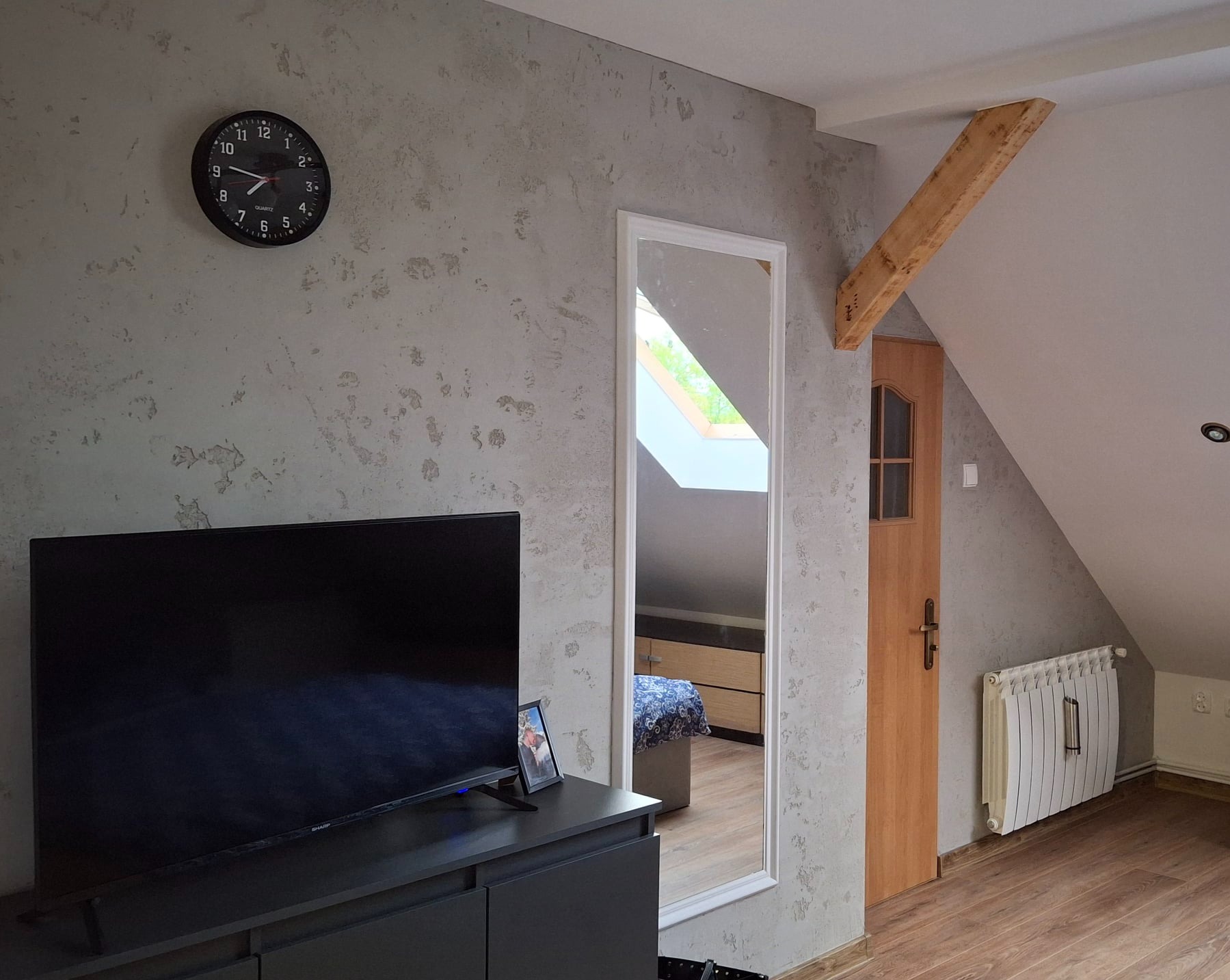 Pokój na poddaszu z szarą ścianą o fakturze betonu, czarnym zegarem i telewizorem. W lustrze odbija się fragment sypialni. Widoczna drewniana belka i drzwi.