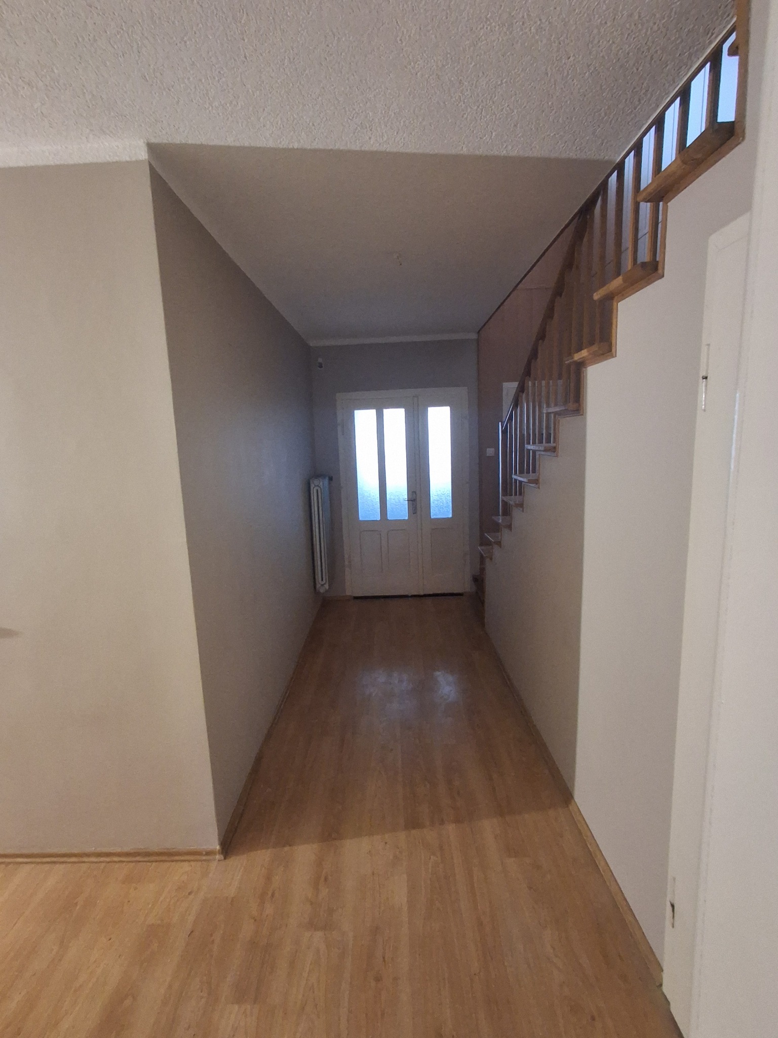 Jasne wnętrze przedpokoju z drewnianą podłogą, schodami i białymi drzwiami wejściowymi. Widoczny grzejnik i neutralne kolory ścian.