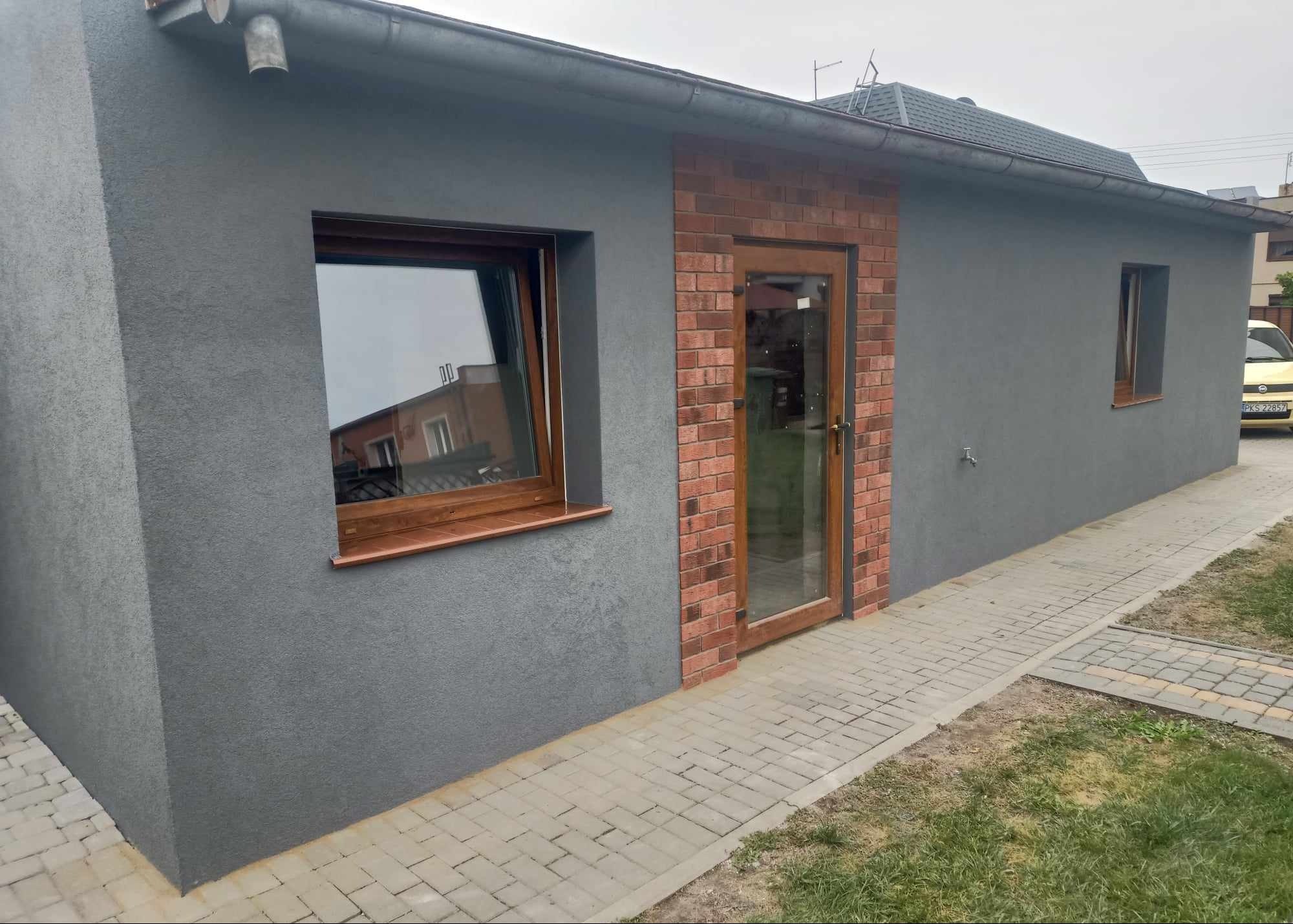 Szary budynek z oknem i drzwiami obramowanymi cegłą, widok z zewnątrz, kostka brukowa, trawa. Elewacja o fakturze baranka. Okno z drewnianą ramą.