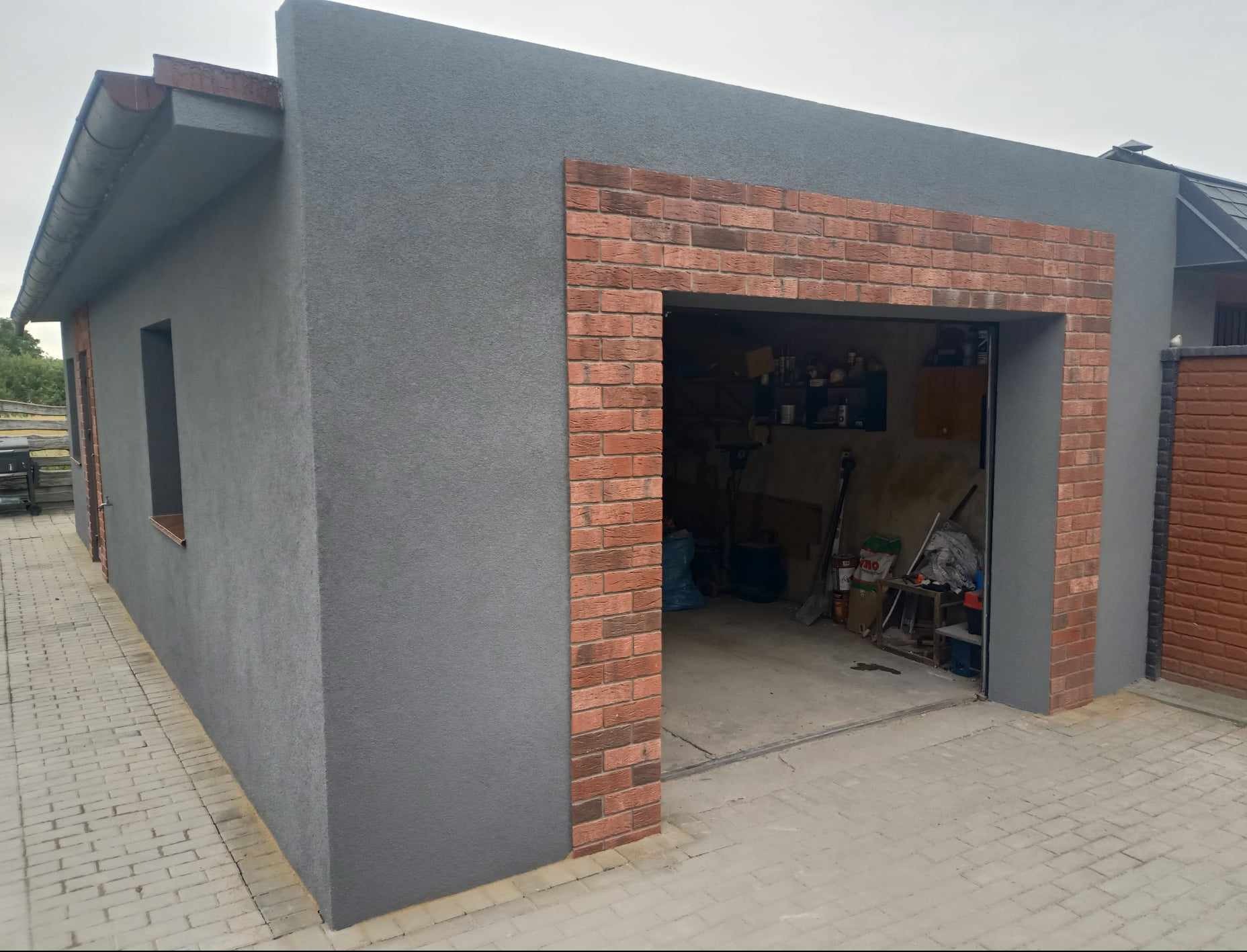 Szary garaż z ceglanym obramowaniem wjazdu, widok z zewnątrz. Wnętrze garażu częściowo widoczne, z widocznymi narzędziami i przedmiotami.