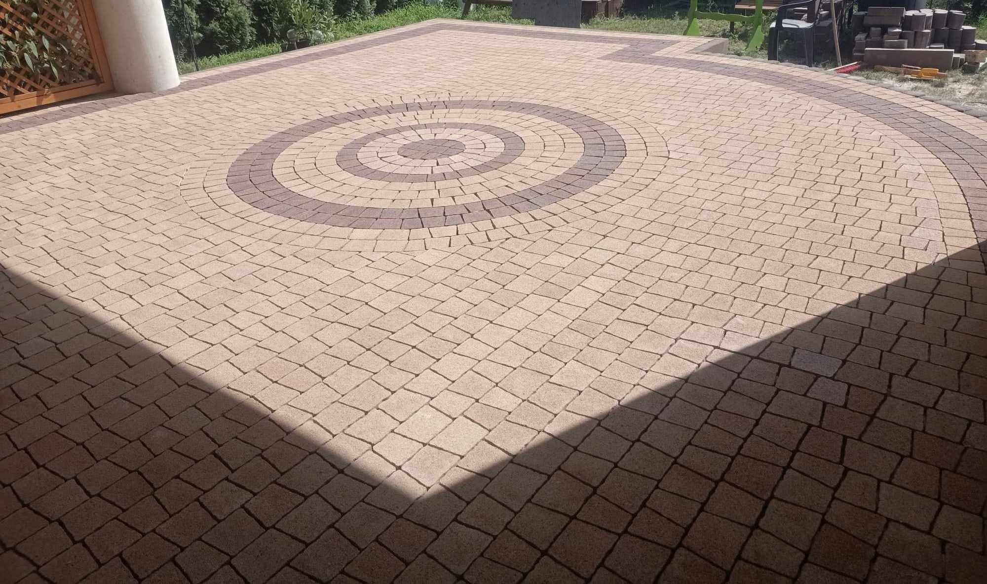 Ułożona kostka brukowa w odcieniach beżu i brązu, tworząca wzór koła. Część powierzchni zacieniona, w tle zieleń. Widoczne elementy drewnianej konstrukcji.