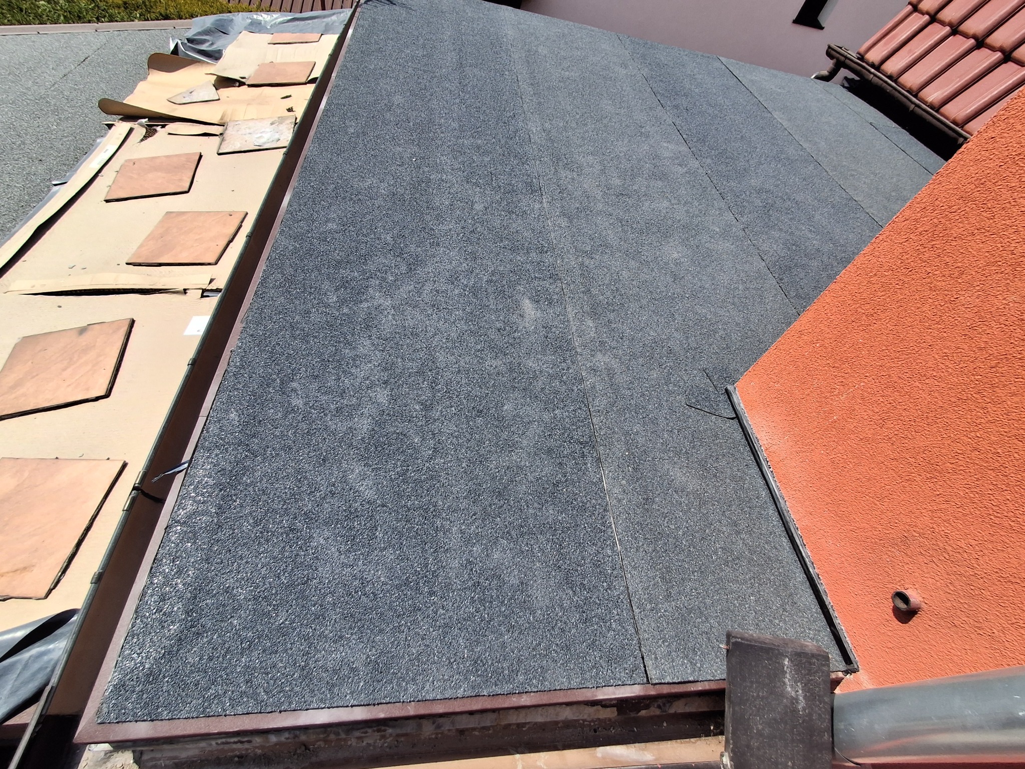 Szczegółowe ujęcie dachu pokrytego papą termozgrzewalną, z widocznymi elementami wykończeniowymi i fragmentem ściany budynku w ciepłym, pomarańczowym odcieniu.