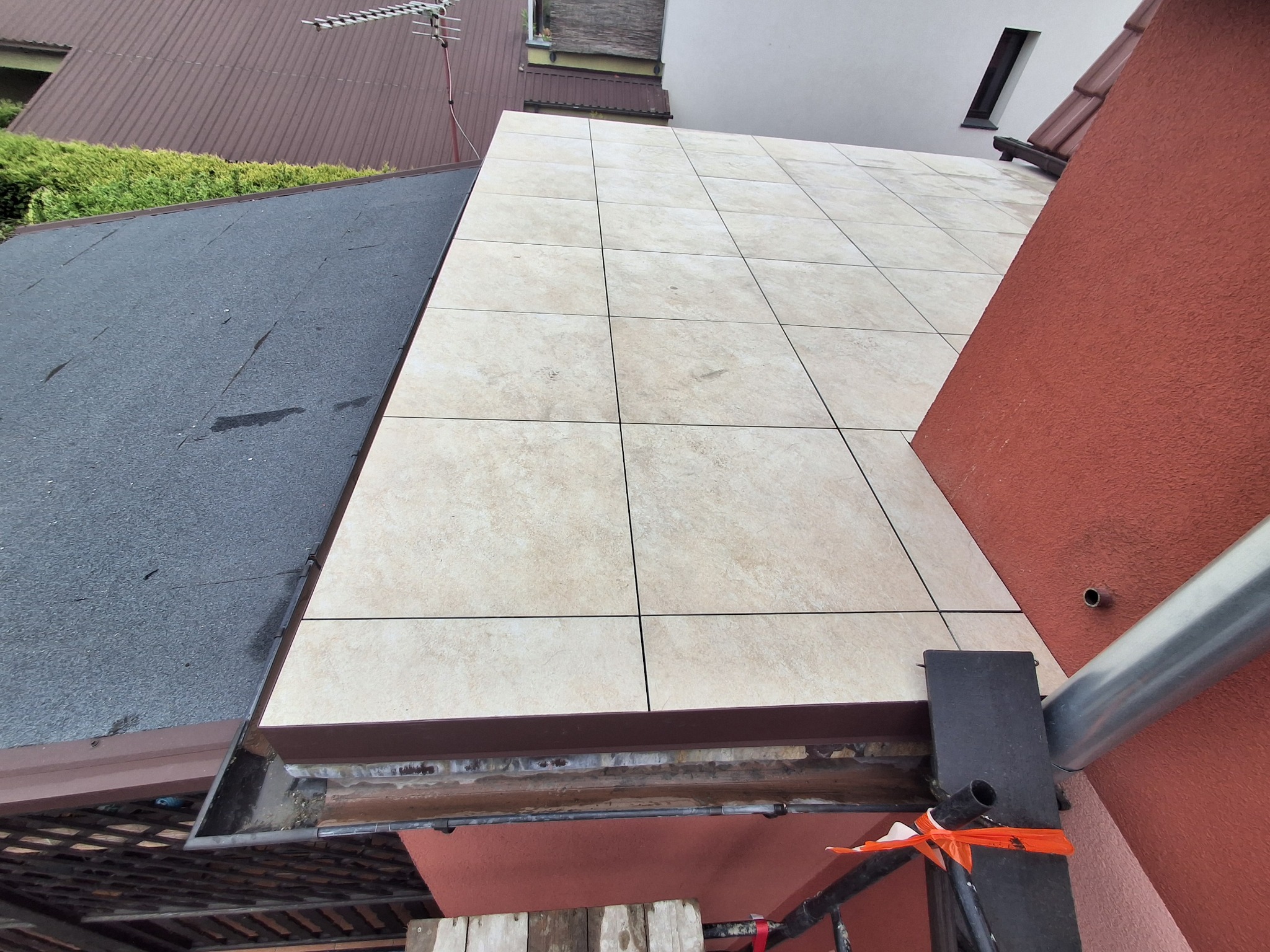 Ułożone płytki na zewnątrz budynku, widok z góry, fragment dachu i ściany. Widoczne rusztowanie i elementy konstrukcyjne. Jasne płytki kontrastują z ciemnym dachem.