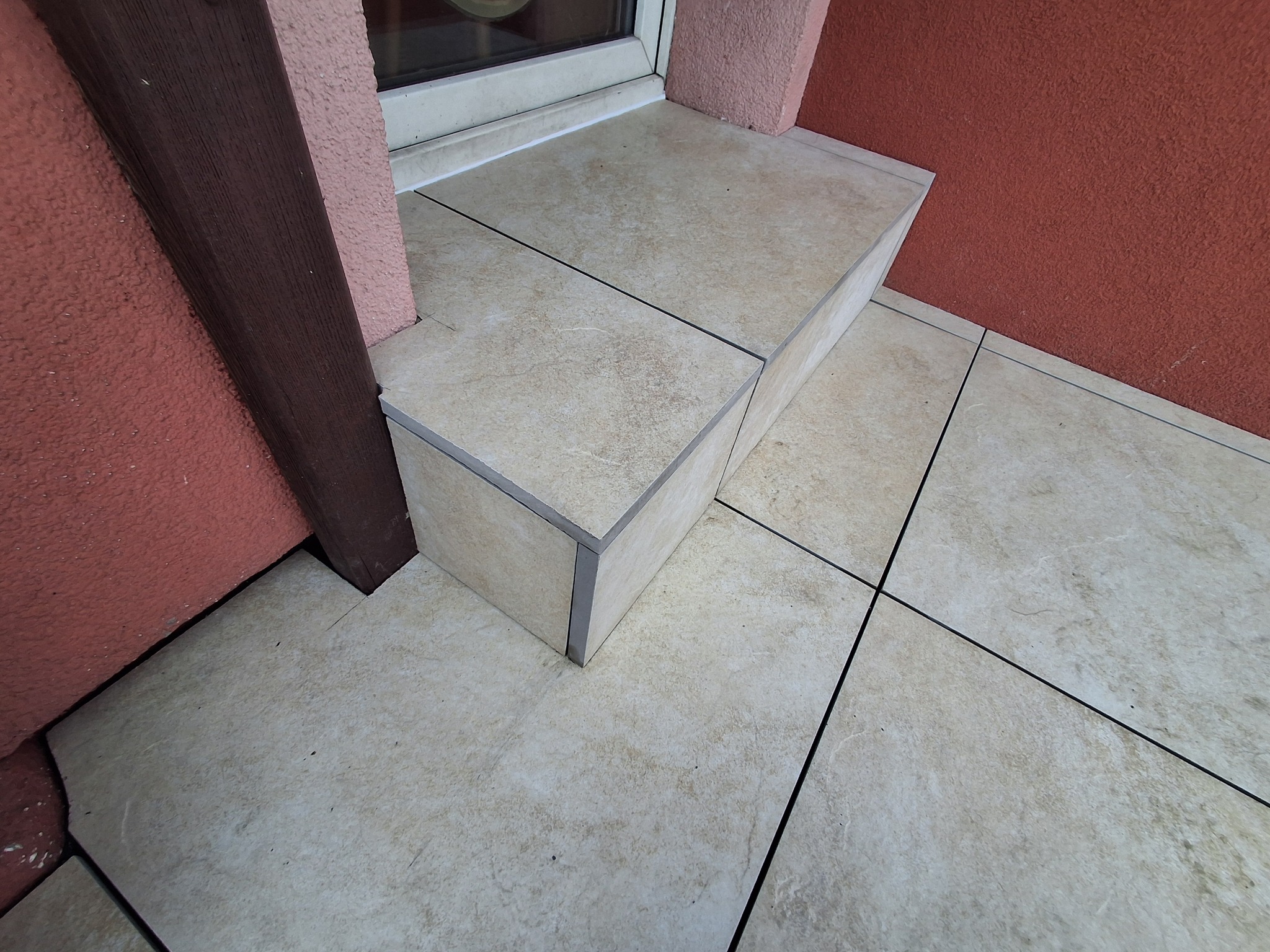 Wykończone płytkami schody wejściowe z beżowych płytek, widoczne fugi i krawędzie wykończeniowe. Ściana o fakturze baranka w odcieniach czerwieni i brązu.