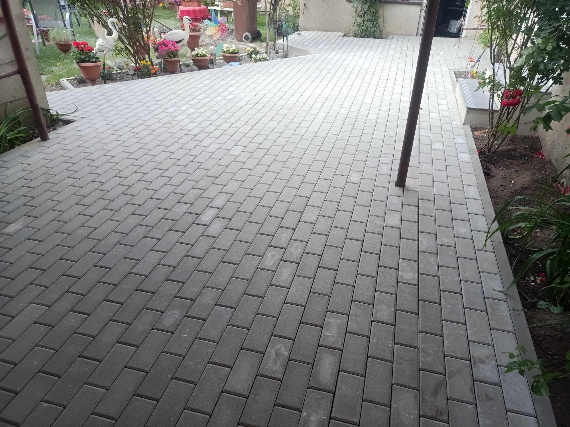 Ułożona szara kostka brukowa na podwórku z elementami zieleni i dekoracjami ogrodowymi. Widok z góry, ukośny kadr.