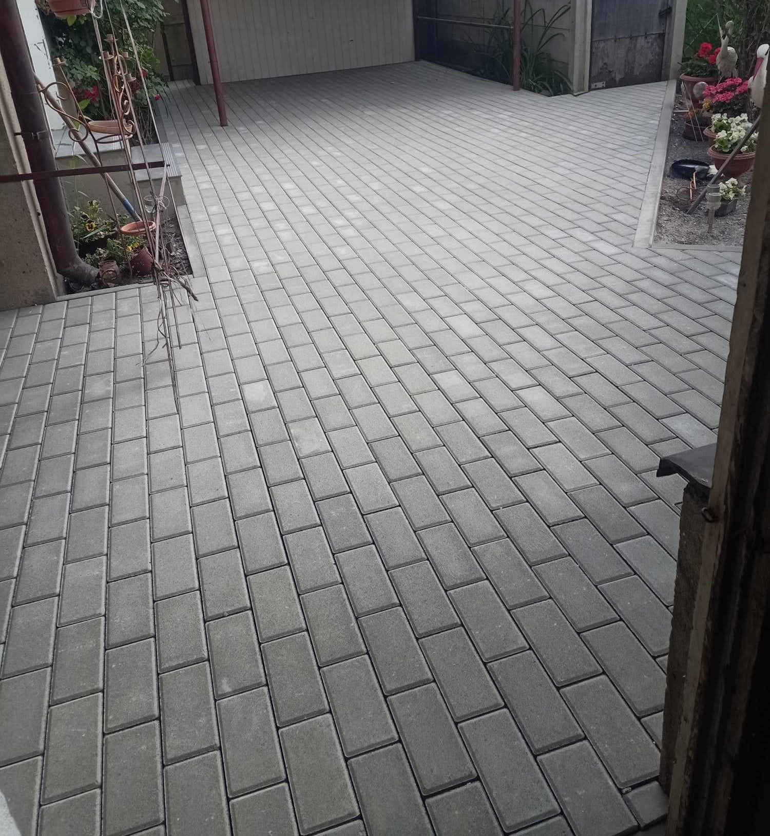 Ułożona szara kostka brukowa na podjeździe, widok z perspektywy drzwi. W tle widoczny garaż i roślinność. Regularny wzór ułożenia kostki.