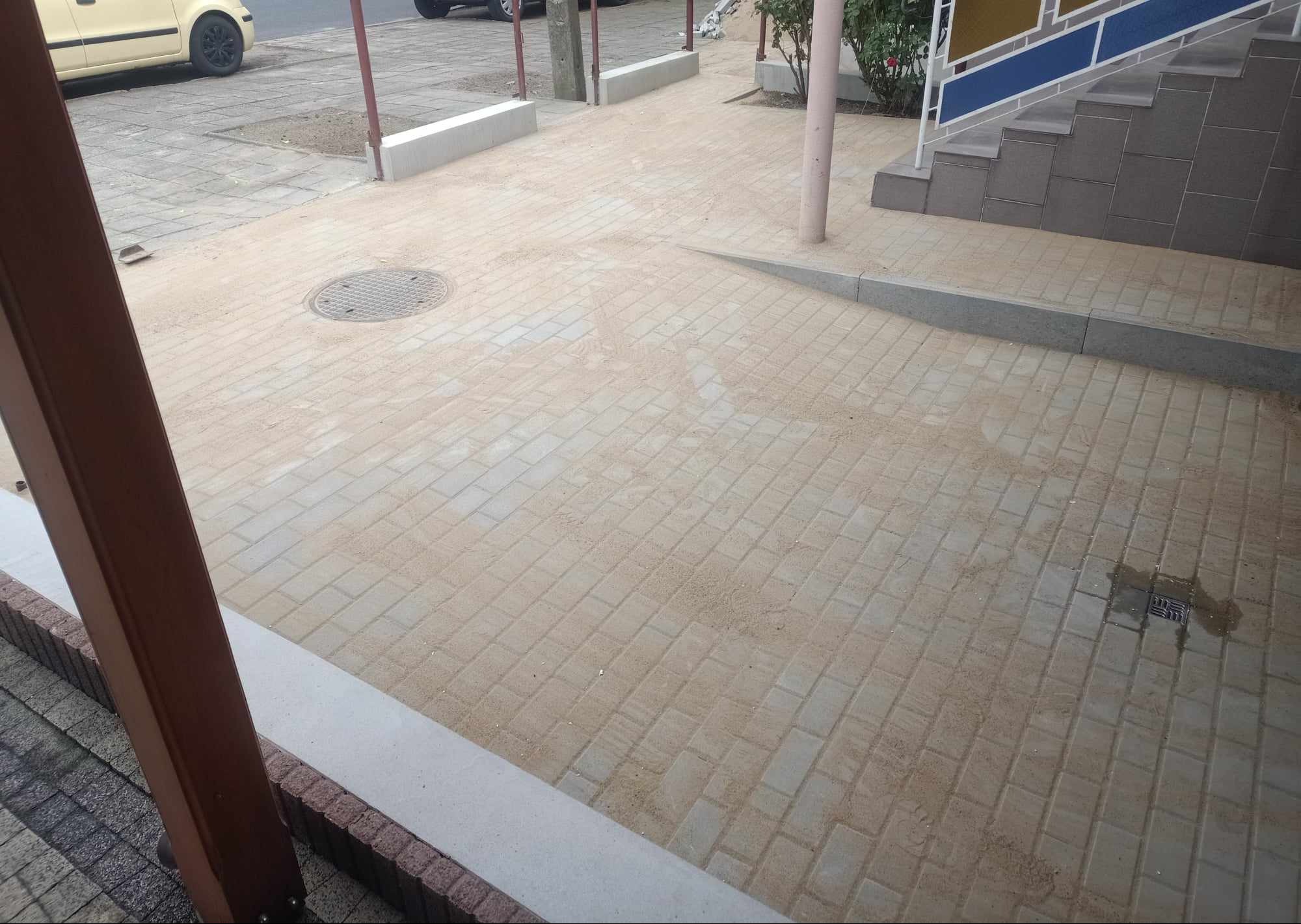 Ułożona kostka brukowa na chodniku, zasypana piaskiem, z widocznym włazem kanalizacyjnym i fragmentem schodów. Widok z góry, lekko z boku.