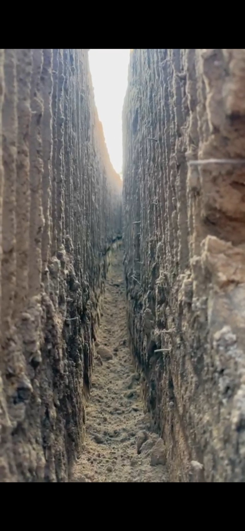 Wąski wykop w ziemi, widok z dołu w górę, ściany wykopu z wyraźną strukturą gleby, na dnie gruz i piasek, w oddali widoczne światło.