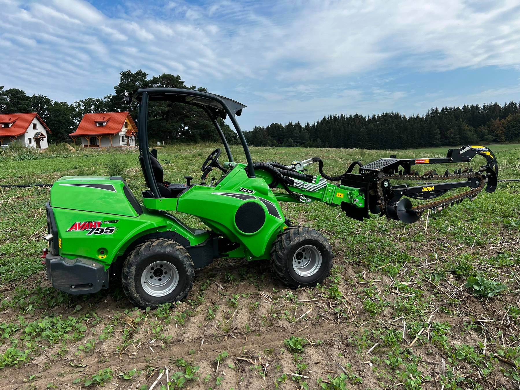 Zielony minikoparko-ładowarka Avant 755 z osprzętem do wykopów, stojąca na polu. W tle widoczne budynki mieszkalne i las, a także niebo z chmurami.
